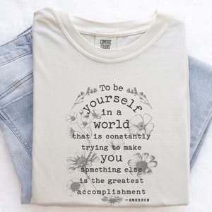 May include: A beige t-shirt with a floral design and the quote "To be yourself in a world that is constantly trying to make you something else is the greatest accomplishment - EMERSON".