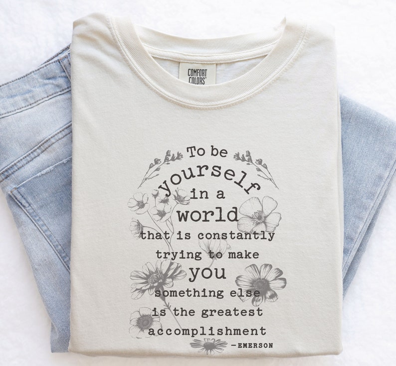 May include: A beige t-shirt with a floral design and the quote "To be yourself in a world that is constantly trying to make you something else is the greatest accomplishment - EMERSON".