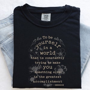 May include: Black t-shirt with a white floral design and the quote "To be yourself in a world that is constantly trying to make you something else is the greatest accomplishment - EMERSON".