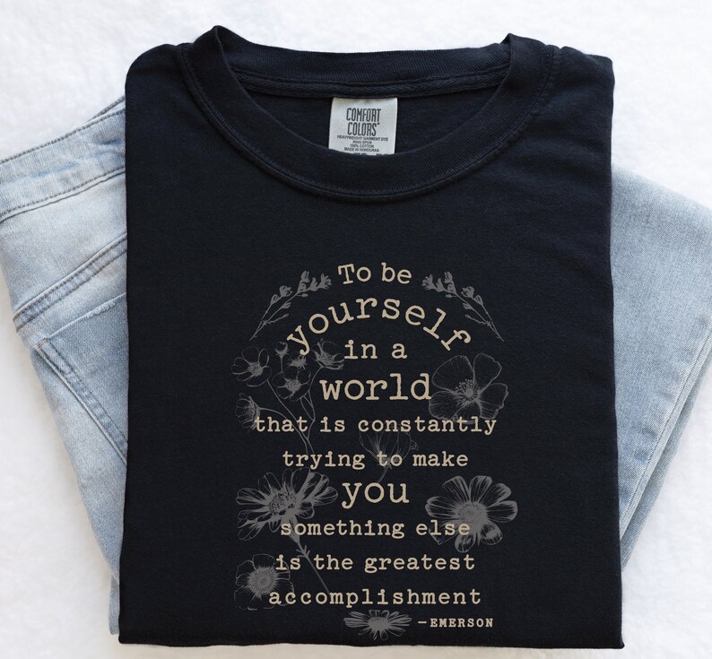 May include: Black t-shirt with a white floral design and the quote "To be yourself in a world that is constantly trying to make you something else is the greatest accomplishment - EMERSON".
