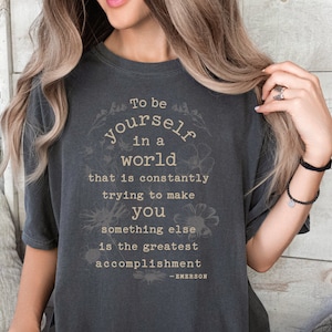 May include: A dark gray t-shirt with a white graphic print that reads "To be yourself in a world that is constantly trying to make you something else is the greatest accomplishment - EMERSON".