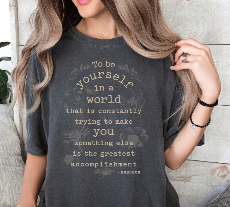 May include: A dark gray t-shirt with a white graphic print that reads "To be yourself in a world that is constantly trying to make you something else is the greatest accomplishment - EMERSON".
