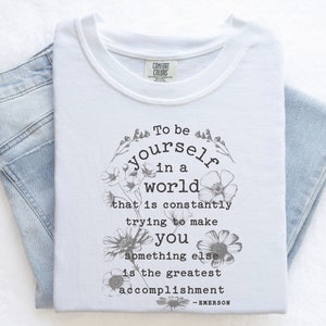 May include: White t-shirt with a floral design and the text "To be yourself in a world that is constantly trying to make you something else is the greatest accomplishment - EMERSON".