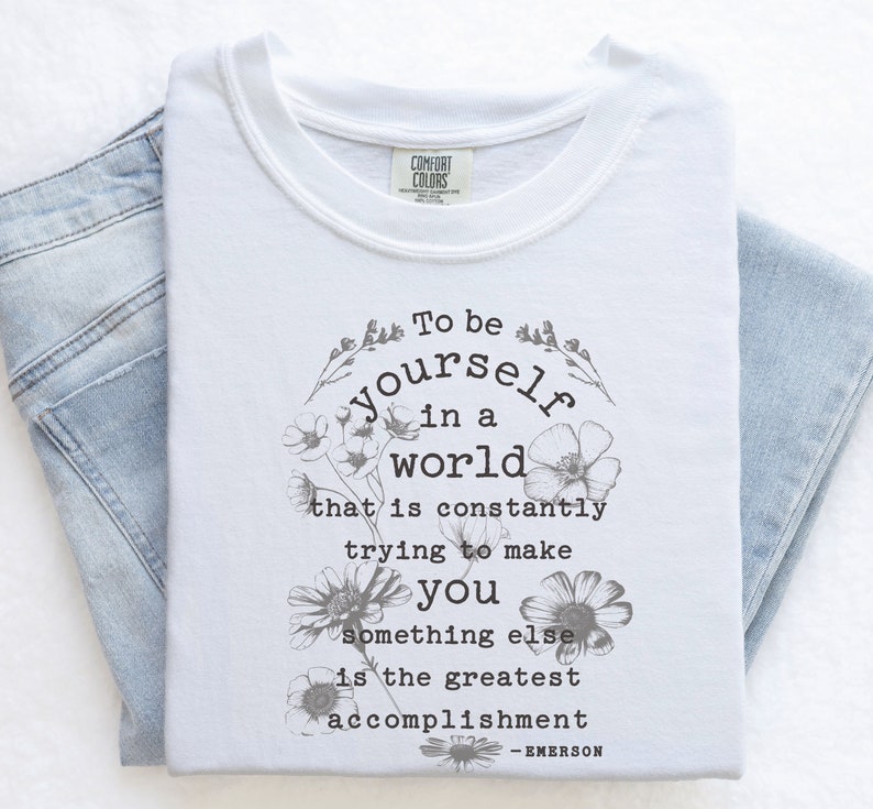 May include: White t-shirt with a floral design and the text "To be yourself in a world that is constantly trying to make you something else is the greatest accomplishment - EMERSON".
