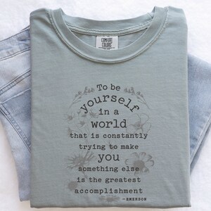 May include: Sage green t-shirt with a floral design and the quote "To be yourself in a world that is constantly trying to make you something else is the greatest accomplishment - EMERSON".
