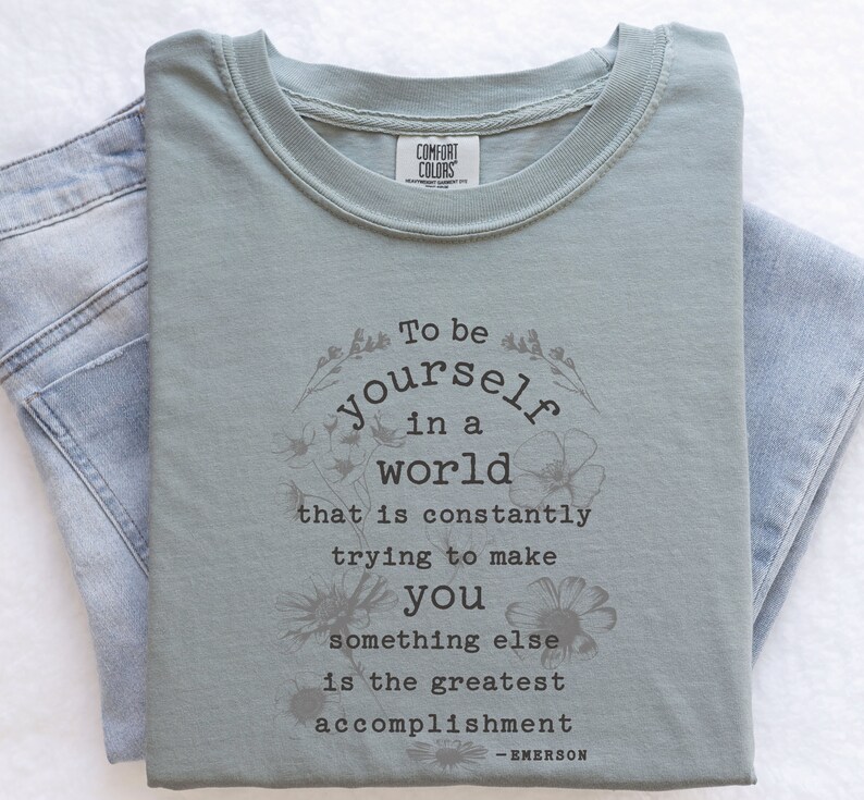 May include: Sage green t-shirt with a floral design and the quote "To be yourself in a world that is constantly trying to make you something else is the greatest accomplishment - EMERSON".