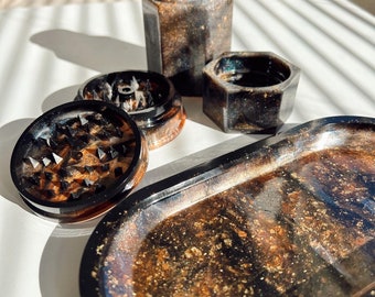 May include: A set of black and gold resin rolling trays, a grinder, and a jar. The trays and grinder have a galaxy-like pattern with gold flecks.