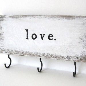 K&ouml;nnte beinhalten: Rechteckiges Holzschild mit dem Wort "love" in schwarzer Schrift. Das Schild ist wei&szlig; gestrichen und hat drei schwarze Metallhaken zum Aufh&auml;ngen von Schl&uuml;sseln oder anderen kleinen Gegenst&auml;nden. Rustikaler Look.