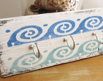 May include: A rustic wooden key holder with a distressed white finish. It features a blue and teal wave pattern and three silver hooks for keys. The key holder is approximately 12 inches long.