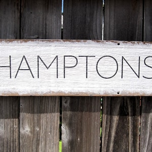May include: A rectangular wooden sign with the word "HAMPTONS" in black lettering. The sign has a white painted surface with distressed edges, mounted on a weathered wooden fence.