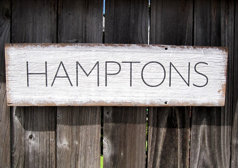 May include: A rectangular wooden sign with the word "HAMPTONS" in black lettering. The sign has a white painted surface with distressed edges, mounted on a weathered wooden fence.