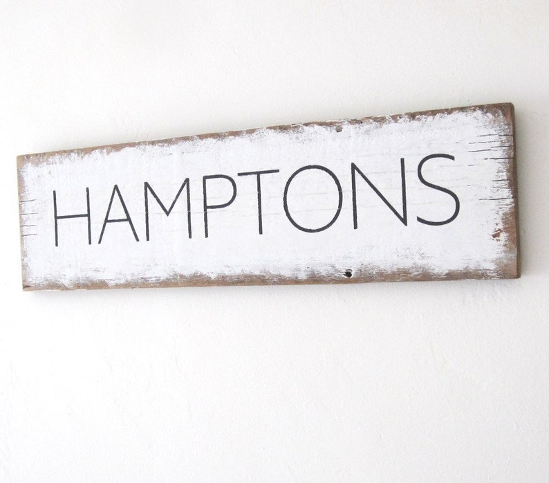 May include: A rectangular wooden sign with the word "HAMPTONS" in black capital letters. The sign has a white painted surface with distressed edges, revealing the natural wood underneath. The sign is mounted on a white wall.