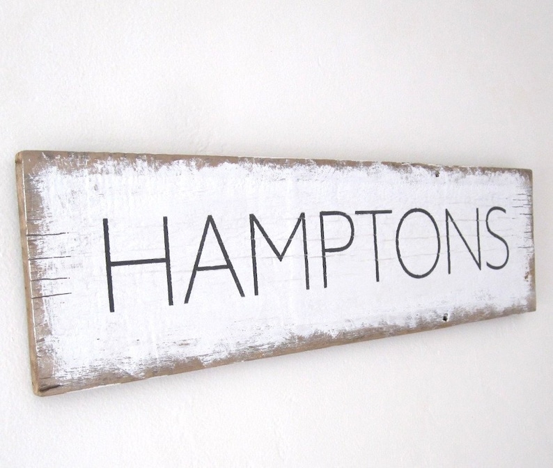 May include: A rectangular wooden sign with the word "HAMPTONS" in black, sans-serif font. The sign has a distressed, rustic look with white paint over the wood and visible wood grain. The edges of the sign are a natural wood color.