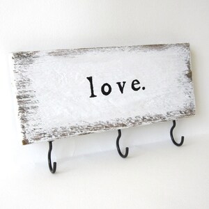 K&ouml;nnte beinhalten: Ein rechteckiges Holzschild mit wei&szlig;er, verwitterter Lackierung. Das Wort "love." ist in Schwarz gedruckt. Drei schwarze Metallhaken sind an der Unterkante angebracht, geeignet zum Aufh&auml;ngen von Schl&uuml;sseln oder anderen kleinen Gegenst&auml;nden.