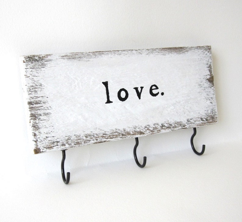 K&ouml;nnte beinhalten: Ein rechteckiges Holzschild mit wei&szlig;er, verwitterter Lackierung. Das Wort "love." ist in Schwarz gedruckt. Drei schwarze Metallhaken sind an der Unterkante angebracht, geeignet zum Aufh&auml;ngen von Schl&uuml;sseln oder anderen kleinen Gegenst&auml;nden.