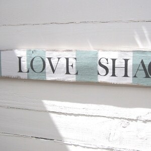 May include: A rectangular wooden sign with the words "THE LOVE SHACK" in black letters. The sign has a distressed, rustic look with alternating white and light blue stripes. The sign is mounted on a white wooden wall.
