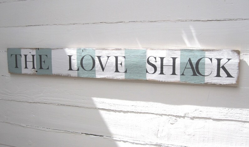 May include: A rectangular wooden sign with the words "THE LOVE SHACK" in black letters. The sign has a distressed, rustic look with alternating white and light blue stripes. The sign is mounted on a white wooden wall.