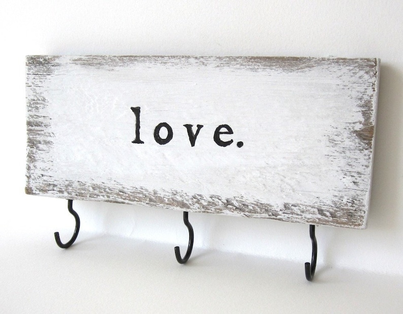 K&ouml;nnte beinhalten: Rechteckiges Holzschild mit dem Wort "love" in schwarzer Schrift. Das Schild ist wei&szlig; gestrichen und hat drei schwarze Metallhaken zum Aufh&auml;ngen von Schl&uuml;sseln oder anderen kleinen Gegenst&auml;nden. Rustikaler Look.