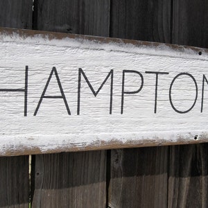 May include: A rustic wooden sign with the word "HAMPTONS" in black lettering. The sign has a distressed white painted background and a natural wood frame, mounted on a weathered wooden fence.