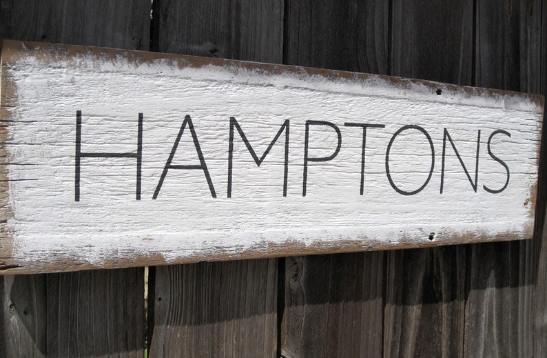 May include: A rustic wooden sign with the word "HAMPTONS" in black lettering. The sign has a distressed white painted background and a natural wood frame, mounted on a weathered wooden fence.