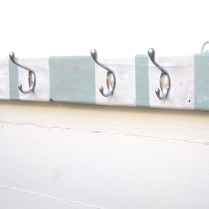 May include: A wooden coat rack with four silver metal hooks. The rack is painted white with sections of light blue-green paint. A metal hanger is attached to the back for mounting. This item is designed for hanging coats, hats, or other items.