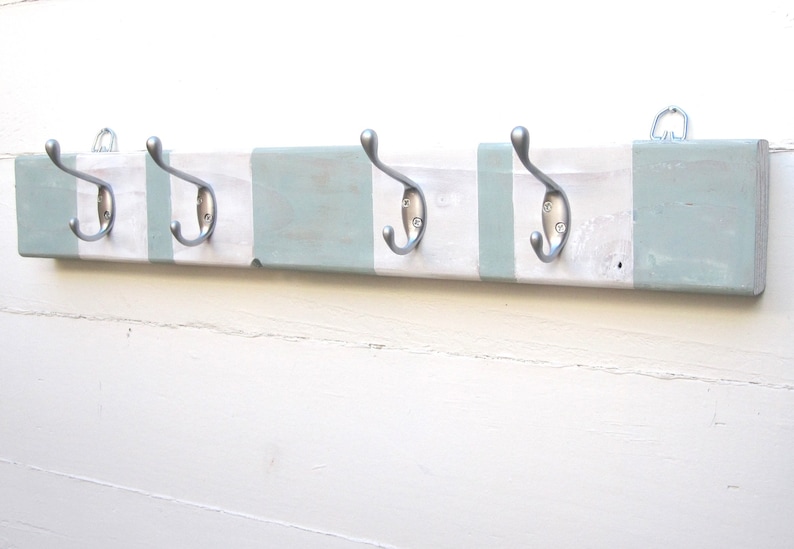 May include: A wooden coat rack with four silver metal hooks. The rack is painted white with sections of light blue-green paint. A metal hanger is attached to the back for mounting. This item is designed for hanging coats, hats, or other items.