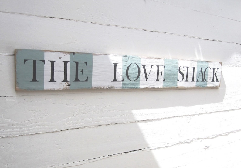 May include: A rectangular wooden sign with the words "THE LOVE SHACK" in black lettering. The sign has a distressed, rustic look with alternating stripes of white and light blue. The sign is mounted on a white, textured wall.