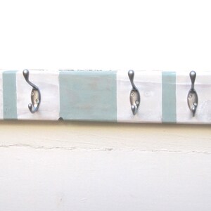 May include: A wooden coat rack with five metal hooks. The rack is painted in alternating stripes of white and light blue. Two metal loops are attached to the top for hanging. The hooks are silver-colored.