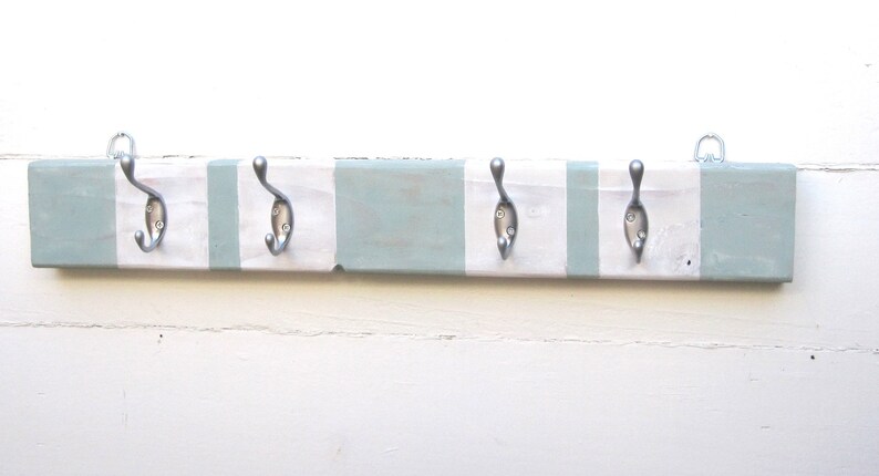 May include: A wooden coat rack with five metal hooks. The rack is painted in alternating stripes of white and light blue. Two metal loops are attached to the top for hanging. The hooks are silver-colored.