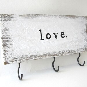 K&ouml;nnte beinhalten: Ein rechteckiger Schl&uuml;sselhalter aus Holz, wei&szlig; gestrichen mit dem schwarzen Wort "love". Er hat drei schwarze Metallhaken. Das Holz hat ein verwittertes Aussehen. Der Schl&uuml;sselhalter ist rechteckig.