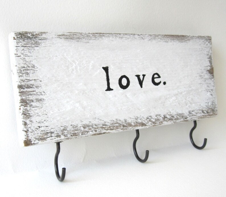 K&ouml;nnte beinhalten: Ein rechteckiger Schl&uuml;sselhalter aus Holz, wei&szlig; gestrichen mit dem schwarzen Wort "love". Er hat drei schwarze Metallhaken. Das Holz hat ein verwittertes Aussehen. Der Schl&uuml;sselhalter ist rechteckig.