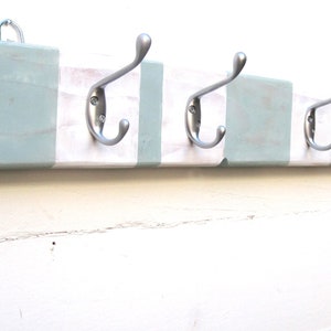 May include: A wooden coat rack with four silver metal hooks. The rack is painted white with light blue stripes. A metal loop is attached to the top for hanging. The design is rustic and suitable for a hallway or entryway.