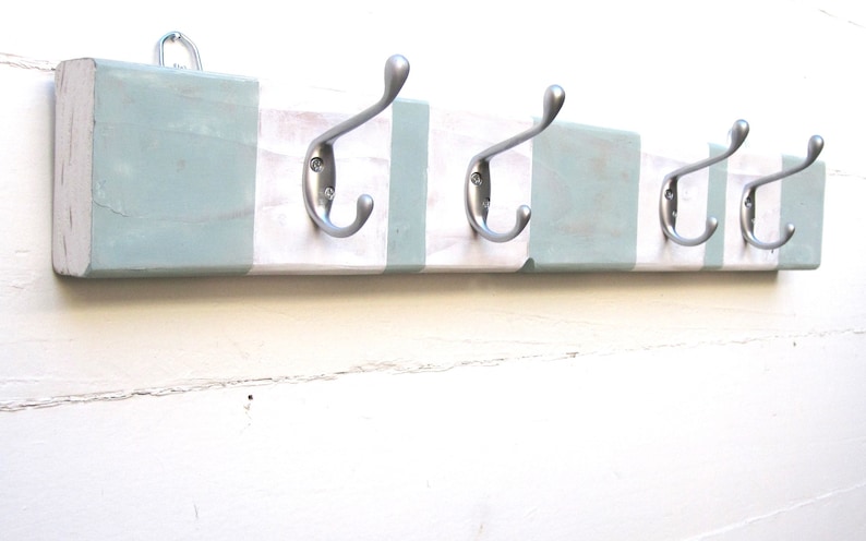 May include: A wooden coat rack with four silver metal hooks. The rack is painted white with light blue stripes. A metal loop is attached to the top for hanging. The design is rustic and suitable for a hallway or entryway.