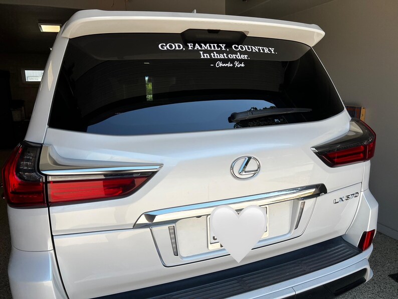 May include: White Lexus SUV with a rear window decal. The decal reads "GOD, FAMILY, COUNTRY. In that order." and is attributed to Charlie Kirk. The vehicle has chrome accents and red taillights.