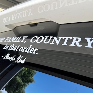 May include: White vinyl decal with the text "GOD, FAMILY, COUNTRY. In that order." and the signature "-Charlie Kirk." The decal is applied to a vehicle's rear window, with the sky visible in the background.