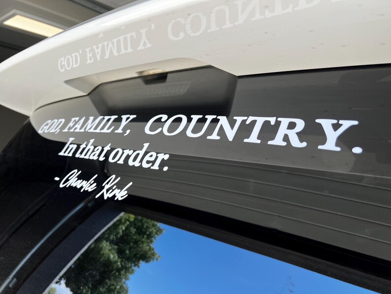 May include: White vinyl decal with the text "GOD, FAMILY, COUNTRY. In that order." and the signature "-Charlie Kirk." The decal is applied to a vehicle's rear window, with the sky visible in the background.