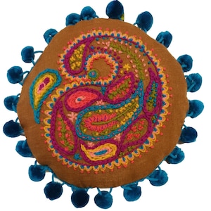 May include: A round brown cushion with a colorful paisley design embroidered on the front. The cushion is surrounded by a blue pom-pom trim.