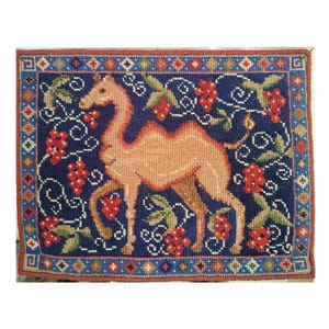 May include: A rectangular needlepoint tapestry depicting a camel in tan hues set against a dark blue backdrop. The design incorporates red grapes, green foliage, and a decorative border with geometric motifs. The tapestry is framed with a brown border.