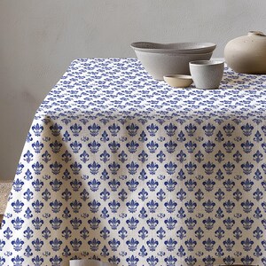 May include: A white tablecloth with a repeating blue fleur-de-lis pattern. The table is set with gray ceramic bowls, a small cup, and a vase. The tablecloth covers the table and drapes over the edge.