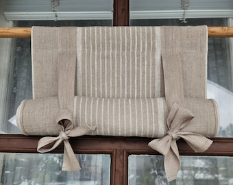 Puede incluir: Una cortina de ventana de lino beige con rayas blancas, enrollada y atada con lazos de tela. La cortina está colgada de una varilla de madera y está delante de una ventana con vista a la nieve.