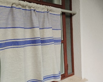 May include: A linen curtain with horizontal blue stripes against a neutral background hangs in a window. The curtain is partially drawn, revealing a wooden window frame and a white wall. The fabric has a textured appearance.