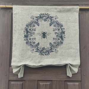 May include: A beige linen curtain with a blue floral wreath and a bee design. The wreath is made of flowers, leaves, and vines. The bee is in the centre of the wreath and has black and white wings.