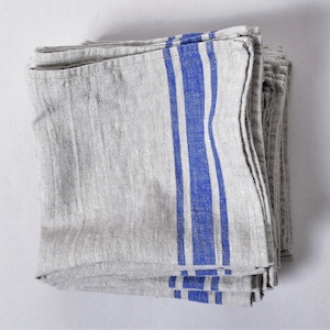 May include: A pile of folded linen napkins in a light grey hue, featuring bold vertical blue stripes. The fabric has a textured finish, suggesting a natural weave.
