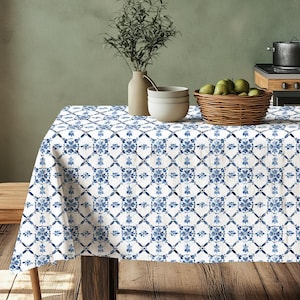 May include: White tablecloth with a blue and white tile pattern. The tablecloth is laid on a wooden table with a vase of greenery, bowls, and a basket of fruit.