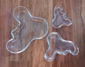 May include: Three clear glass serving dishes in organic shapes. The largest dish is shaped like a teardrop with a rounded bottom. The two smaller dishes are shaped like a kidney bean.