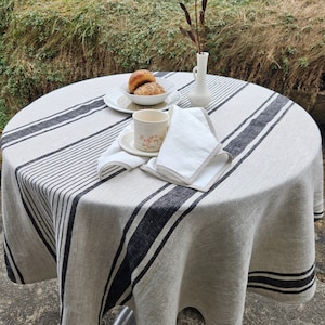 Puede incluir: Un mantel redondo de lino blanco con rayas negras. El mantel está puesto sobre una mesa con una taza y un platillo de cerámica blanca, una servilleta blanca y un plato pequeño con dos rollos.