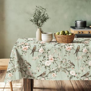 Può includere: Una tovaglia verde con un motivo floreale con rose bianche e uccelli marroni. La tovaglia è appoggiata su un tavolo di legno con un vaso di rami verdi e una ciotola di frutta.