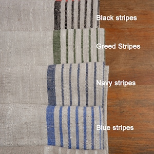 May include: A set of eight linen tea towels with different coloured stripes. The towels are arranged in a stack, with the top towel having burgundy stripes, the next towel having red stripes, the next towel having black stripes, the next towel having green stripes, the next towel having navy stripes, the next towel having blue stripes, and the bottom towel having natural stripes.