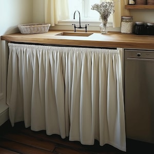 Puede incluir: Una cortina de cocina blanca con un dobladillo con volantes cuelga frente a un lavavajillas de acero inoxidable. La cortina está hecha de una tela ligera y tiene un diseño simple.