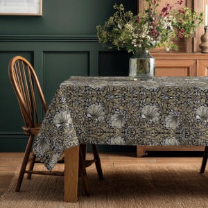 May include: A rectangular tablecloth featuring a dense, repeating botanical pattern of large, stylised cream-coloured flowers, dark green leaves, and intricate brown vines on a black background, draped over a wooden table with a wooden chair.